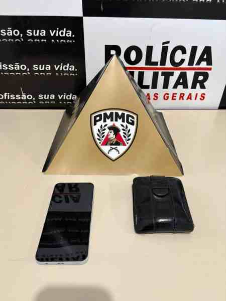Uma pirâmide dourada com o emblema da PMMG, acompanhada de um celular e uma carteira preta sobre uma mesa.