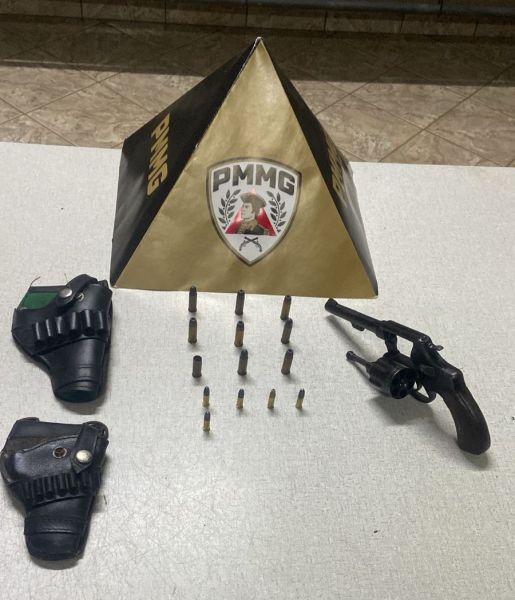 Uma pirâmide com o logo da PMMG, duas coldres de arma, um revólver e vários cartuchos dispostos em linha sobre uma mesa.