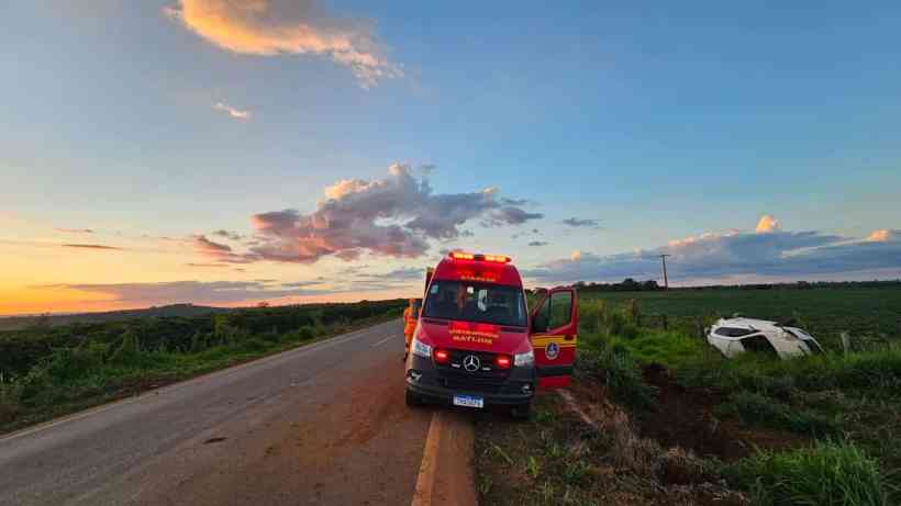 Ambulância da corporação de resgate parada ao lado da estrada, com as luzes de emergência ligadas, enquanto um carro acidentado está fora da pista ao fundo, sob um céu colorido de pôr do sol.