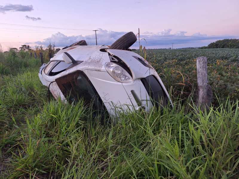 Um carro branco tombado em um campo cercado por grama alta e vegetação.