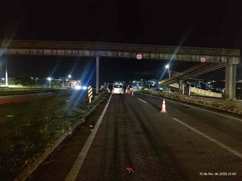 Foto de uma rodovia à noite, com sinalização de trânsito e cones de alerta, abaixo de uma ponte.