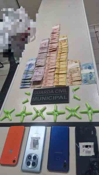 Mesa com notas de dinheiro empilhadas em forma de pirâmide, placa da Guarda Civil Municipal e vários telefones celulares dispostos ao lado.