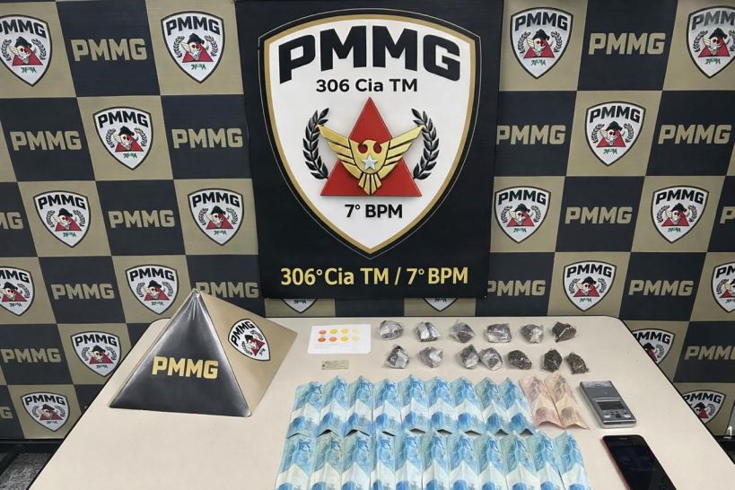 Mesa com uma pirâmide metálica com o símbolo PMMG, dinheiro, porções de substâncias em sacos plásticos e um celular, com um fundo de plaquinhas da PMMG.