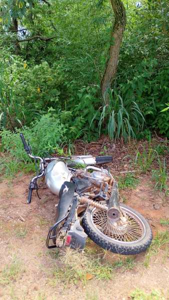 Motocicleta caída em uma área arbustiva, coberta de sujeira e com vegetação ao fundo.