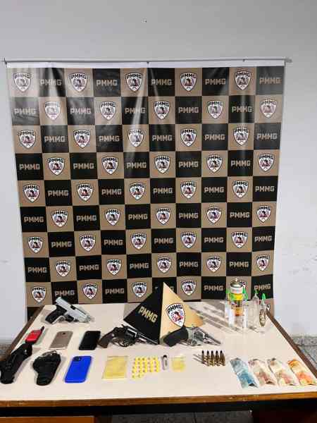 Mesa com armas, munições e objetos diversos, incluindo drogas e celulares, em frente a um fundo com logotipos da PMMG.