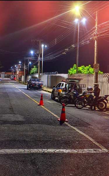 Uma rua à noite com iluminação pública, contendo cones de sinalização, um veículo da polícia e uma motocicleta parada.