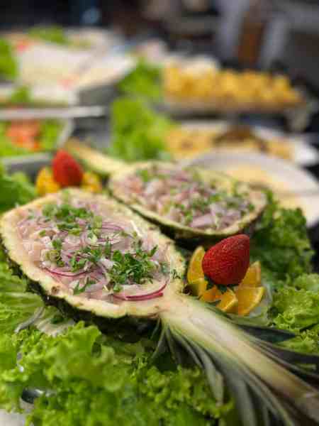 Prato decorativo com abacaxi cortado ao meio, recheado com peixe, cebola roxa e ervas, rodeado por folhas de alface e fatias de frutas como morango e laranja.