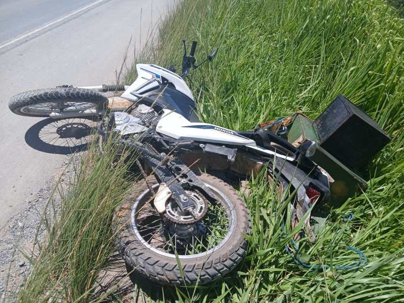 Motocicleta caída em um gramado ao lado da estrada, com pneus danificados e caixa ao fundo.