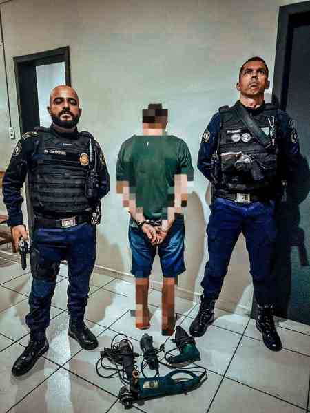 Dois policiais em pé ao lado de um homem detido, que está com as mãos para trás. No chão, há ferramentas de perfuração.