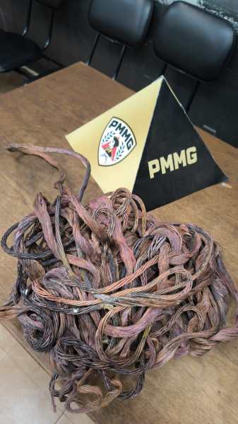 Um emaranhado de fios de metal escuros e marrons sobre uma mesa de madeira, com um objeto piramidal ao fundo exibindo o logotipo da PMMG.