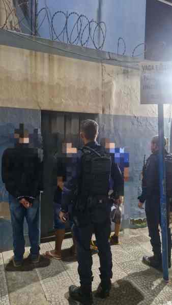 Policiais em operação, abordando suspeitos em frente a um muro com arame farpado.