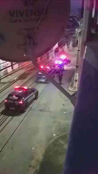 Cena noturna mostrando veículos da polícia com luzes acesas em uma rua, com um policial em pé enquanto outros carros estão estacionados.