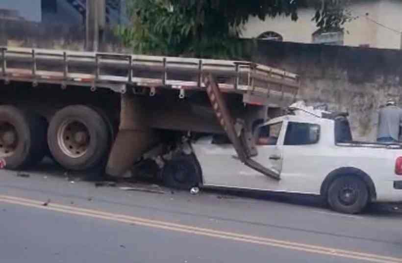 Caminhão tombado sobre um carro branco em uma rua, com danos visíveis e pessoas ao fundo avaliando a situação.
