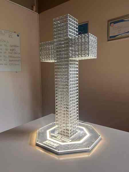 Escultura de uma cruz iluminada, feita de material transparente com padrão de malha, sobre uma base octogonal iluminada, em ambiente interno.