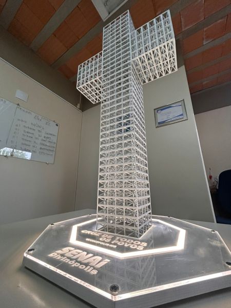 Escultura em forma de cruz feita de material transparente, iluminada na base, com uma placa em baixo que contém o nome 'SENAI' e a palavra 'Dinatorpólis'.