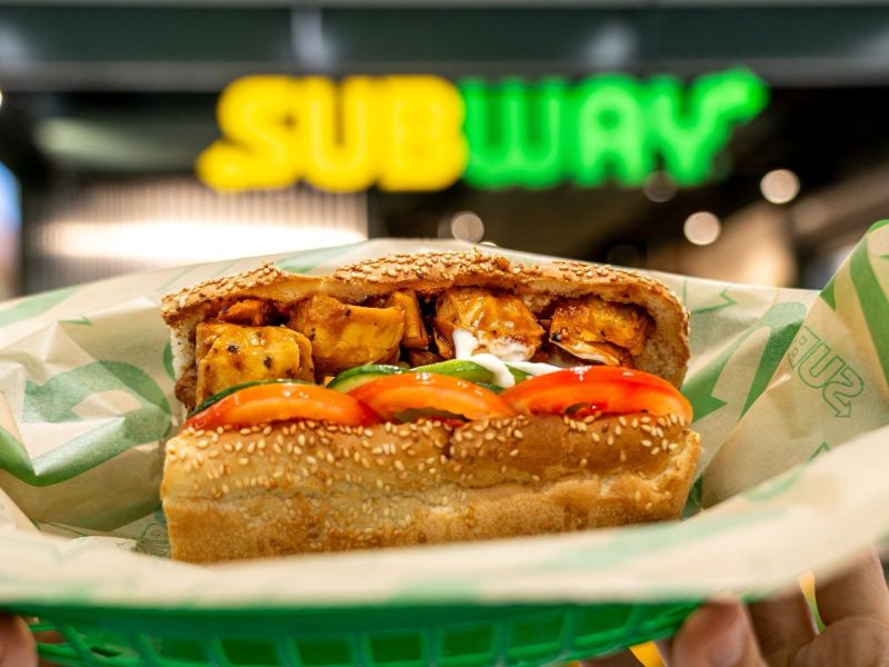 A close-up of a sesame seed sandwich filled with chunks of grilled chicken, sliced tomatoes, green peppers, and a creamy sauce, held in a green basket with a blurred Subway logo in the background.