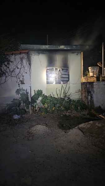 Uma pequena casa à noite, com um lado queimado e uma janela coberta por grades. Vegetação ao redor da casa e uma área de terra ao primeiro plano.