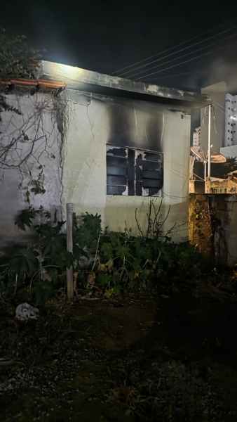 Casa parcialmente queimada à noite, com janelas danificadas e vegetação ao redor.