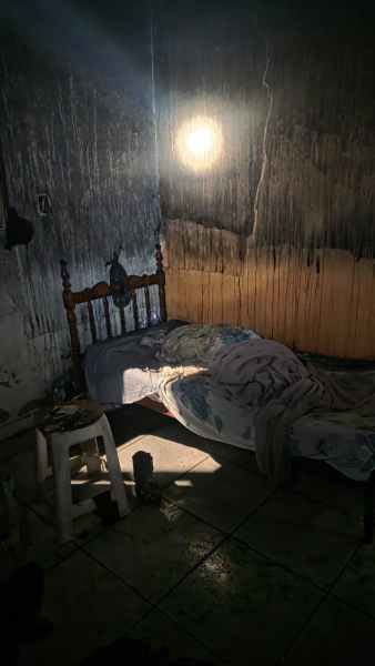 Quarto escuro com uma cama desfeita, iluminado por uma luz suave, paredes manchadas com marcas de desgaste e um assento branco ao lado.