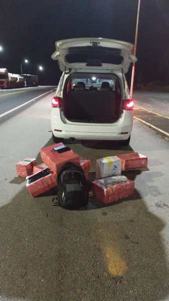 Um carro branco com a porta traseira aberta, exibindo pacotes vermelhos e uma mochila no chão ao lado da estrada durante a noite.